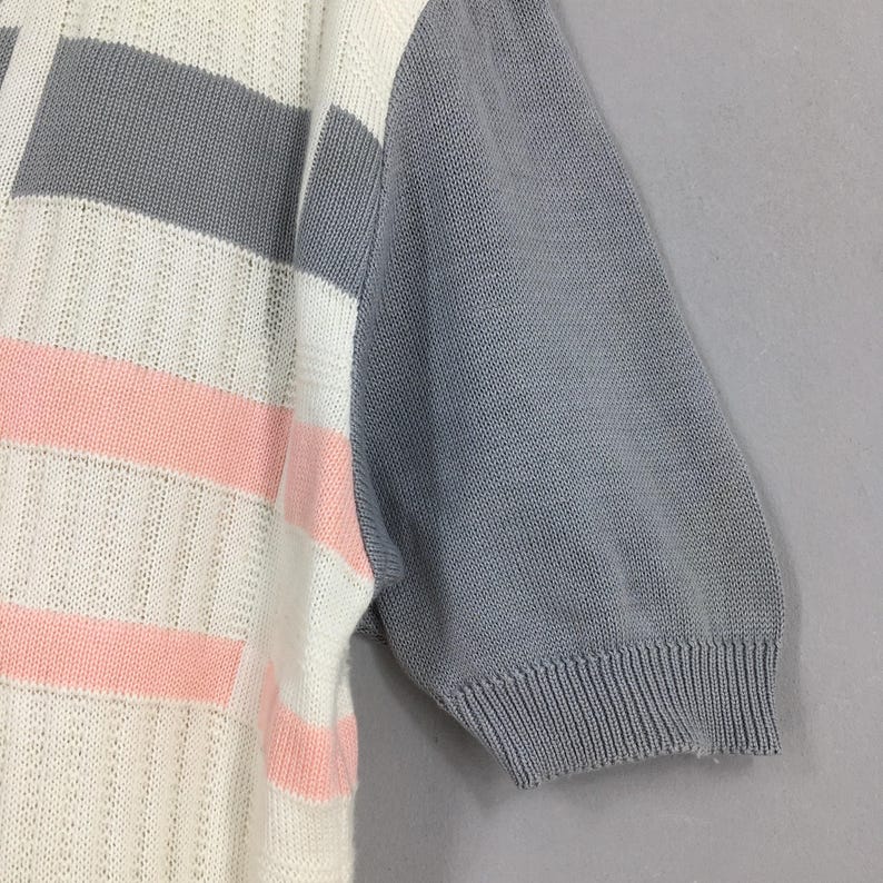 May include: Close-up of a short-sleeved knit top. The top features a color block design with white, gray, and pink horizontal stripes on the front. The sleeve is solid gray. The knit fabric has a textured appearance.