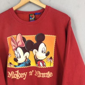 May include: Red sweatshirt featuring a graphic of Mickey and Minnie Mouse. The graphic includes the text "Mickey n' Minnie" in a vintage style font. The sweatshirt has a crew neck and long sleeves. The tag reads "Mickey Unlimited".