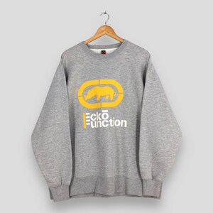 May include: A heather gray crewneck sweatshirt featuring a yellow graphic of a rhinoceros inside an oval outline. Below the graphic, the text Ecko Function is printed in white and yellow. The garment has long sleeves and a ribbed hem and cuffs.