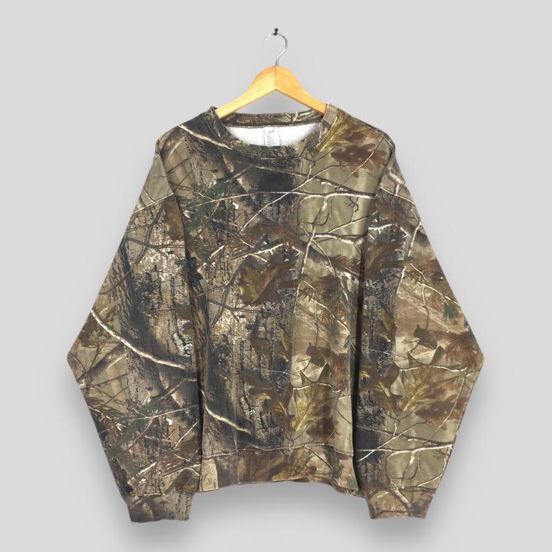 May include: A camouflage-patterned sweatshirt hangs on a wooden hanger. The sweatshirt features a brown, green, and tan woodland camouflage design. The long-sleeved garment has a crew neck and ribbed cuffs and hem. The background is a solid light gray.