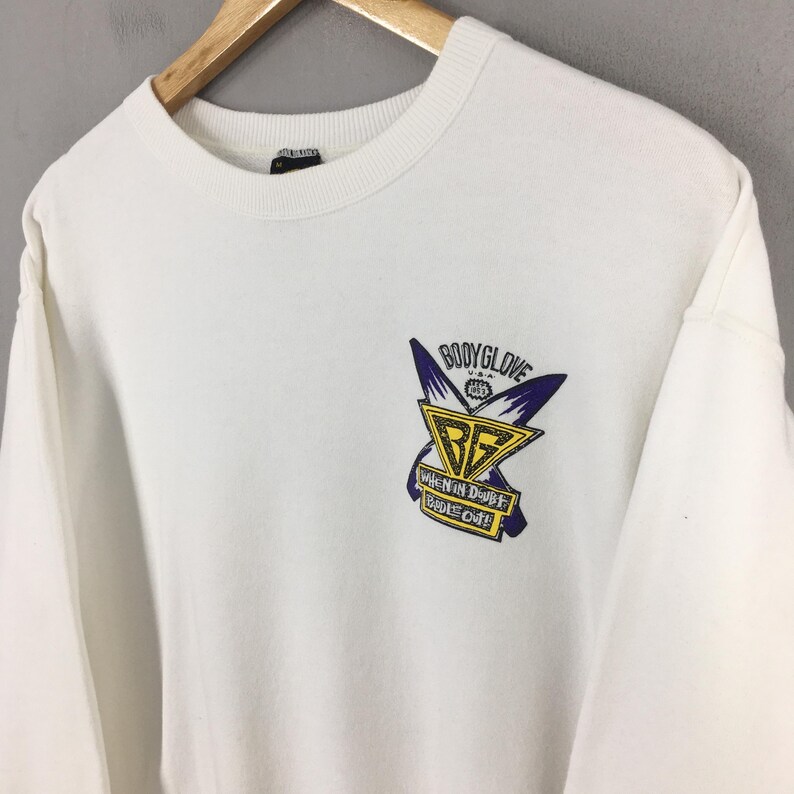 Op de afbeelding: Witte crewneck sweatshirt met een Body Glove USA logo op de borst. Het logo heeft een gele en zwarte driehoek met de letters "BG" erin, en de tekst "WHEN IN DOUBT PADDLE OUT!"