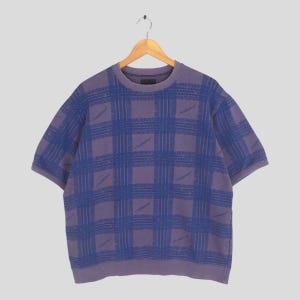 May include: A short-sleeved, crew neck shirt in a purple hue, featuring a blue plaid pattern. The shirt is hanging on a wooden hanger against a light gray background. The plaid design is made up of repeating geometric shapes.