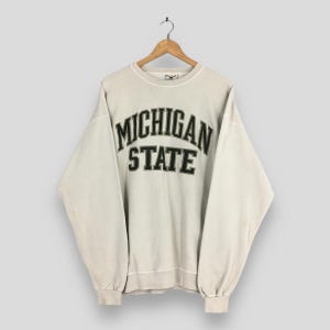 Puede incluir: Una sudadera beige claro con las palabras "MICHIGAN STATE" en letras negras y verdes. La sudadera está colgada de una percha de madera.