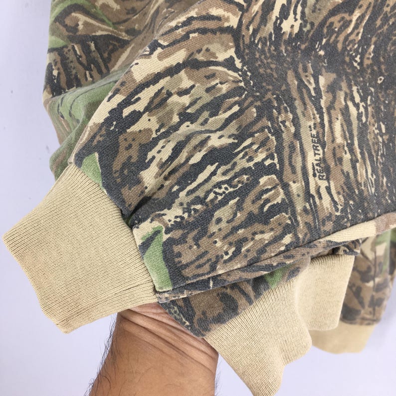 Op de afbeelding: Close-up van een camouflage-gepatroneerde sweatshirtmouw. De stof heeft een mix van bruine, groene en bruine kleuren, met een geribbelde bruine manchet. Het woord "Realtree" is op de mouw gedrukt.