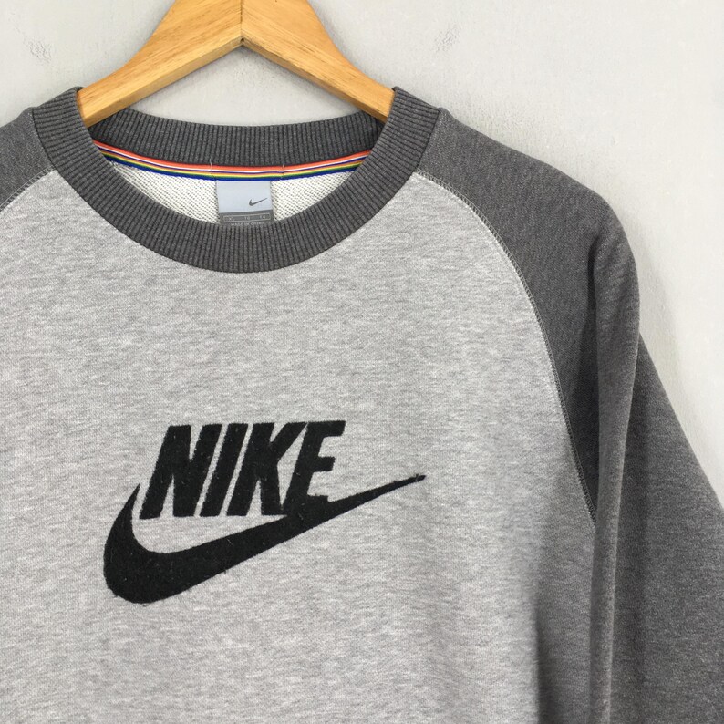 K&ouml;nnte beinhalten: Graues Nike Sweatshirt mit Rundhalsausschnitt und Raglan&auml;rmeln. Das Nike-Logo ist in Schwarz auf der Vorderseite zu sehen. Der Kragen hat ein gestreiftes Detail.