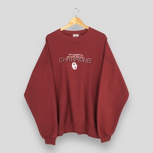 May include: A maroon Lee sweatshirt with the text "OKLAHOMA 2000 NATIONAL FOOTBALL CHAMPIONS" in white and silver lettering. The letter "O" is also featured. The sweatshirt is hanging on a wooden hanger.