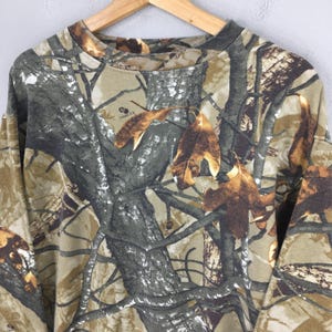 May include: A long-sleeved camouflage shirt with a crew neck. The shirt features a pattern of brown, green, and tan colors, resembling a forest scene with branches and leaves. The shirt is hanging on a wooden hanger.