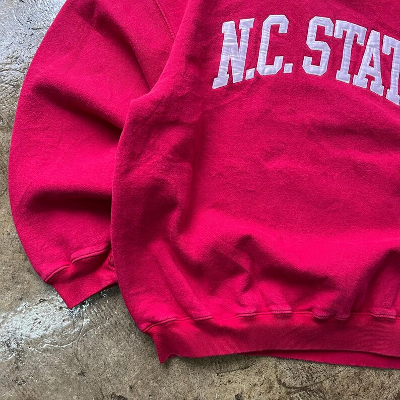 May include: A vibrant pink sweatshirt with the text "N.C. STATE" in white block letters across the top. The sweatshirt has a crew neck and ribbed cuffs and hem. The fabric appears soft and slightly worn.