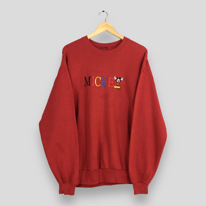 Peut inclure: Un sweat-shirt rouge avec le mot "MICKEY" brod&eacute; en couleurs vives. Une petite image brod&eacute;e de Mickey Mouse est pr&eacute;sente. Le sweat-shirt est suspendu &agrave; un cintre en bois.