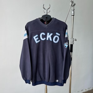 May include: Navy blue V-neck sweatshirt with the word "ECKO" in light blue across the chest. The sleeves have white and light blue stripes. A small patch is on the right sleeve. The number "19" is at the bottom.