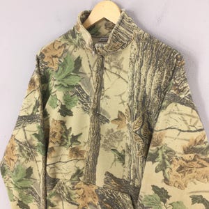 Puede incluir: Sudadera de camuflaje con cremallera parcial y bolsillo tipo canguro. El dise&ntilde;o presenta una mezcla de colores marr&oacute;n, verde y beige, que se asemejan a una escena forestal. La sudadera est&aacute; colgada en una percha de madera.