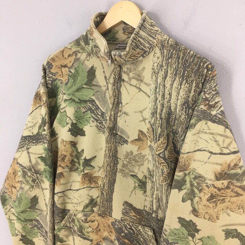 Puede incluir: Sudadera de camuflaje con cremallera parcial y bolsillo tipo canguro. El dise&ntilde;o presenta una mezcla de colores marr&oacute;n, verde y beige, que se asemejan a una escena forestal. La sudadera est&aacute; colgada en una percha de madera.