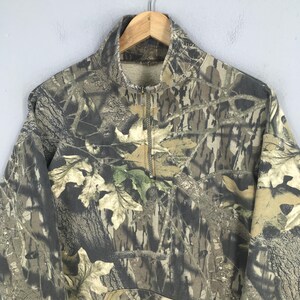 Pode incluir: Camisola de camuflagem com fecho de correr de um quarto. O design apresenta uma mistura de tons castanhos, verdes e bege, imitando um ambiente florestal. A gola &eacute; de cor lisa e a pe&ccedil;a de vestu&aacute;rio est&aacute; pendurada num cabide de madeira.