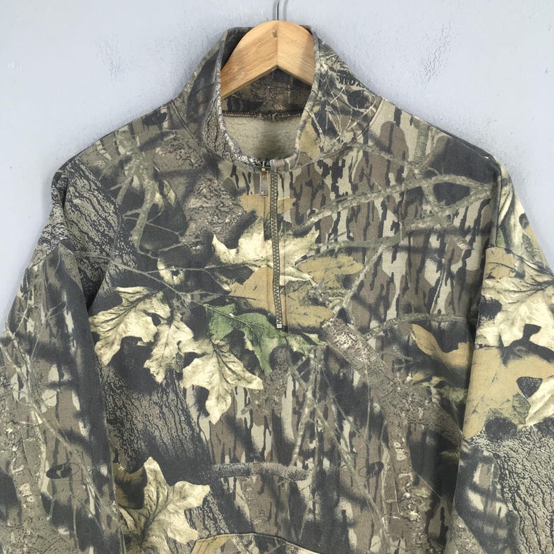 Pode incluir: Camisola de camuflagem com fecho de correr de um quarto. O design apresenta uma mistura de tons castanhos, verdes e bege, imitando um ambiente florestal. A gola &eacute; de cor lisa e a pe&ccedil;a de vestu&aacute;rio est&aacute; pendurada num cabide de madeira.