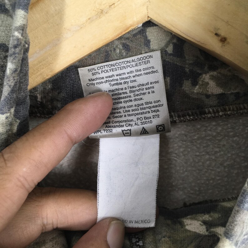 May include: Close-up of a tag attached to a camouflage-patterned garment. The tag includes washing instructions and fabric content: 50% cotton and 50% polyester. The tag also indicates the garment was made in Mexico.