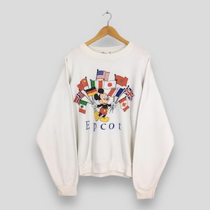 May include: White sweatshirt featuring Mickey Mouse holding flags from various countries. The word "Epcot" is printed in blue below the graphic. The sweatshirt has long sleeves and a crew neck, perfect for casual wear.