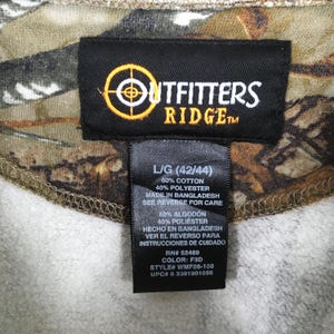 May include: Close-up of a camouflage-patterned garment tag. The tag features the "Outfitters Ridge" logo in white and yellow, with text indicating size L/G (42/44), fabric composition, and care instructions. The tag is black with white text.