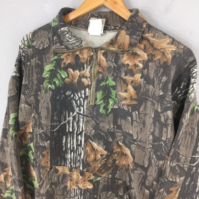 K&ouml;nnte beinhalten: Ein Sweatshirt mit Camouflage-Muster und einem Viertel-Rei&szlig;verschluss. Das Design zeigt Braun-, Gr&uuml;n- und Graut&ouml;ne mit einem Muster aus Bl&auml;ttern und Baumrinde. Der Kragen ist in einem helleren Grauton gehalten.