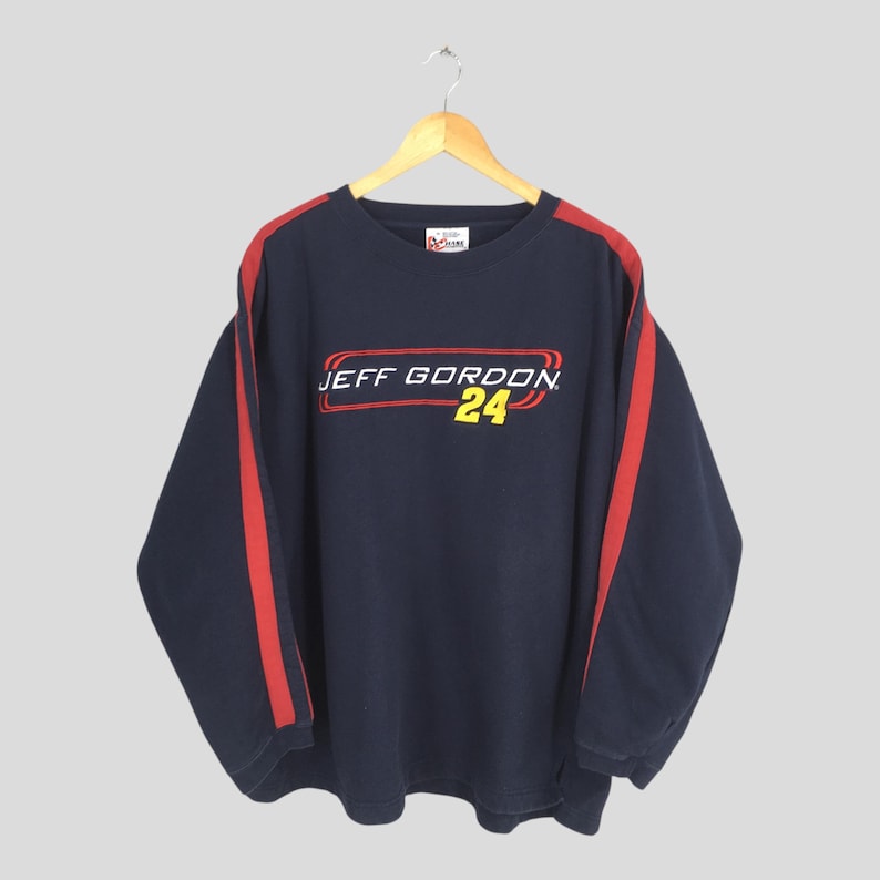 Puede incluir: Sudadera azul marino de manga larga con rayas rojas en las mangas. La parte delantera presenta el texto "JEFF GORDON 24" en blanco, rojo y amarillo. La sudadera est&aacute; colgada de una percha de madera.