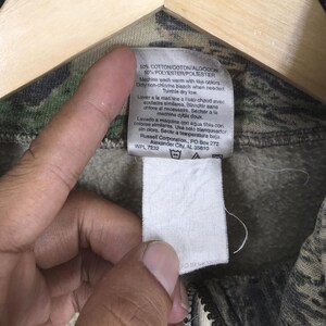Puede incluir: Primer plano de una etiqueta blanca con texto negro adjunta a una prenda con estampado de camuflaje. La etiqueta proporciona detalles de la tela e instrucciones de lavado. La prenda es una mezcla de algod&oacute;n y poli&eacute;ster.