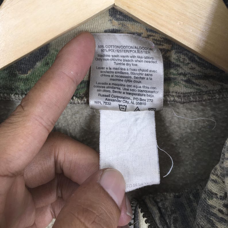 Puede incluir: Primer plano de una etiqueta blanca con texto negro adjunta a una prenda con estampado de camuflaje. La etiqueta proporciona detalles de la tela e instrucciones de lavado. La prenda es una mezcla de algod&oacute;n y poli&eacute;ster.