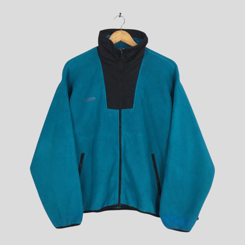May include: Teal fleece jacket with a black collar and zipper. The jacket has a high collar and two front pockets. A small logo is visible on the upper left chest area. The jacket is hanging on a wooden hanger.