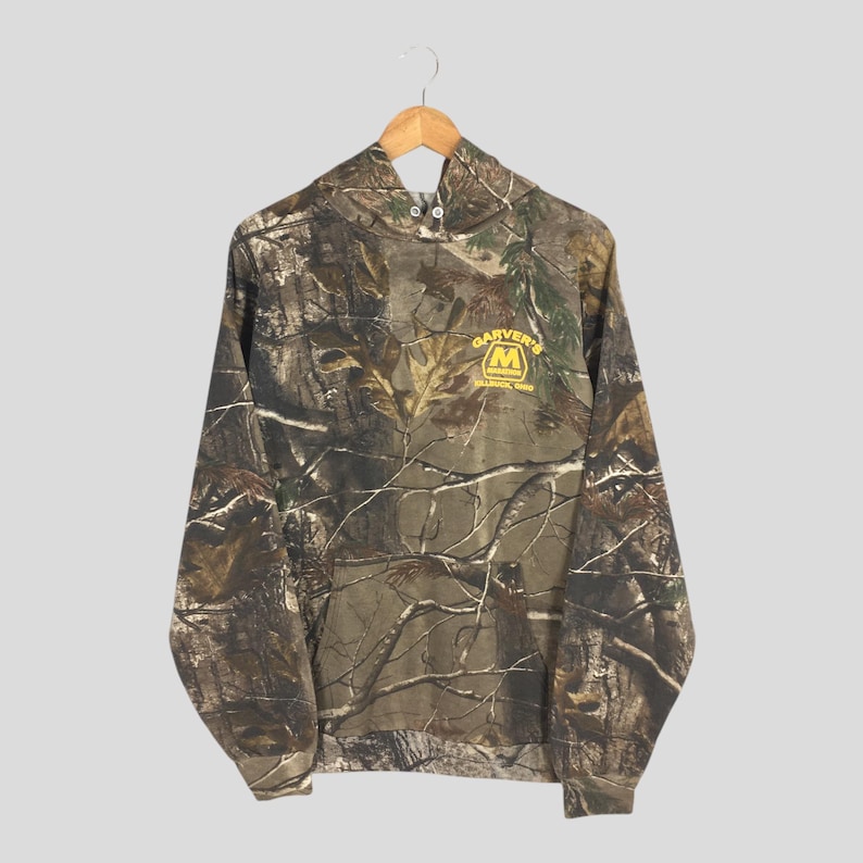 May include: A camouflage-patterned hoodie with a drawstring hood and long sleeves. The hoodie features the text "GARVER'S" above a yellow "M" logo, with "MALVERN, OHIO" below. The garment is hanging on a wooden hanger.