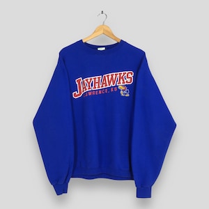 Pode incluir: Uma camisola azul royal com a palavra "JAYHAWKS" em letras vermelhas e brancas. Abaixo da palavra está "LAWRENCE, KS" em letras brancas. Um pequeno logotipo de Jayhawk de desenho animado está no lado direito.
