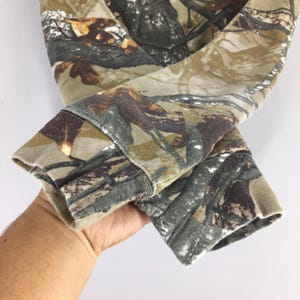 May include: Close-up of a camouflage-patterned long-sleeved garment. The fabric features a blend of brown, green, and tan hues, mimicking a forest environment. The cuffs are visible, showcasing the garment's design and texture.