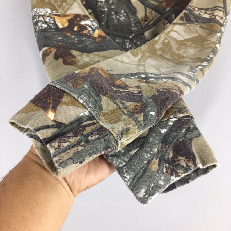 May include: Close-up of a camouflage-patterned long-sleeved garment. The fabric features a blend of brown, green, and tan hues, mimicking a forest environment. The cuffs are visible, showcasing the garment's design and texture.