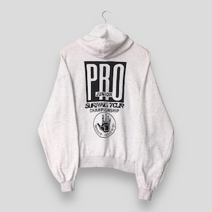 Puede incluir: Sudadera con capucha gris claro con capucha y mangas largas. La parte trasera presenta un cuadrado negro con la palabra "PRO" en su interior, junto con "JUNIOR SURFING TOUR CHAMPIONSHIP" y un logotipo de mano.
