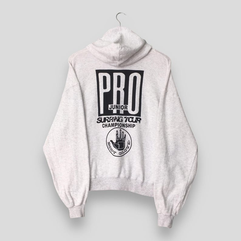 Puede incluir: Sudadera con capucha gris claro con capucha y mangas largas. La parte trasera presenta un cuadrado negro con la palabra "PRO" en su interior, junto con "JUNIOR SURFING TOUR CHAMPIONSHIP" y un logotipo de mano.