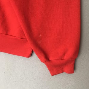 Peut inclure: Un sweat-shirt rouge vif &agrave; manches longues dot&eacute; d'un poignet en tricot c&ocirc;tel&eacute; et d'un ourlet c&ocirc;tel&eacute; assorti au bas. Le tissu pr&eacute;sente une texture rouge unie et lisse, avec une petite tache claire et l&eacute;g&egrave;re visible sur la manche.