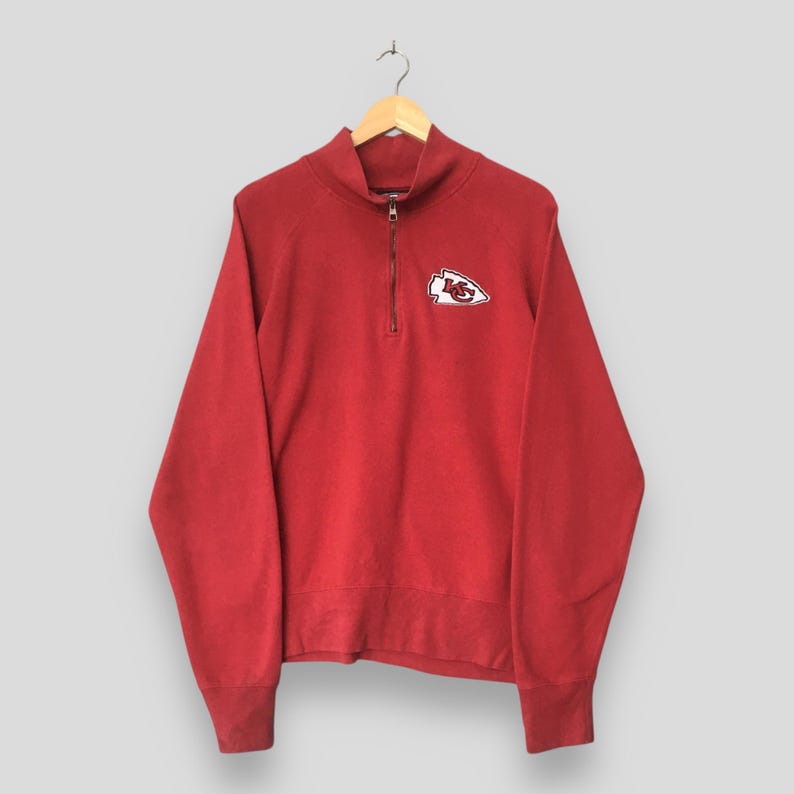 Op de afbeelding: Een rood sweatshirt met lange mouwen en een korte rits, voorzien van een wit en rood Kansas City Chiefs pijlpuntsymbool geborduurd op de linkerborst. Het kledingstuk heeft een hoge kraag en geribbelde boorden aan mouwen en zoom.