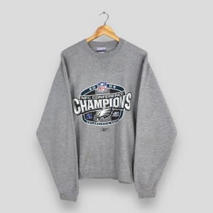 Könnte beinhalten: Ein graumeliertes Langarm-Sweatshirt mit Rundhalsausschnitt und einem Grafikdruck mit der Aufschrift 2004 NFL NFC CONFERENCE CHAMPIONS, dem Logo der Philadelphia Eagles, einem Reebok-Logo und einem kleinen NFL-Wappen auf der Brust.