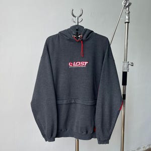Könnte beinhalten: Ein dunkelgrauer Kapuzenpullover mit dem Text "LOST ENTERPRISES" in Rot und Weiß auf der Brust. Der Kapuzenpullover hat eine Kapuze mit Kordelzug und roten Akzenten an den Ärmeln. Der Hoodie hängt an einem Metallbügel.