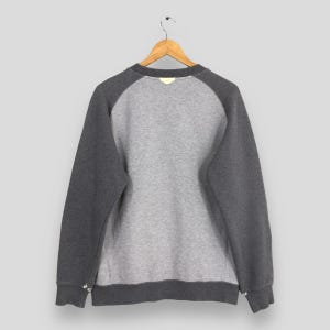 K&ouml;nnte beinhalten: Ein graues Sweatshirt mit Raglan&auml;rmeln, das an einem Holzb&uuml;gel h&auml;ngt. Der K&ouml;rper des Sweatshirts ist hellgrau, mit dunkelgrauen &Auml;rmeln und Bes&auml;tzen. Die R&uuml;ckseite des Kleidungsst&uuml;cks ist sichtbar.