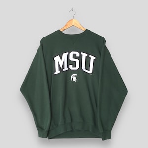 Puede incluir: Una sudadera verde oscuro con las letras blancas "MSU" en letras de bloque, delineadas en negro. Debajo de las letras hay un logotipo de casco espartano blanco. La sudadera está colgada de una percha de madera.