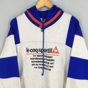 May include: A vintage Le Coq Sportif hoodie in white, blue, and red. The hoodie features the brand's logo and French text. The design includes blue panels on the sleeves and a blue and red striped neckline. The hoodie is hanging on a wooden hanger.