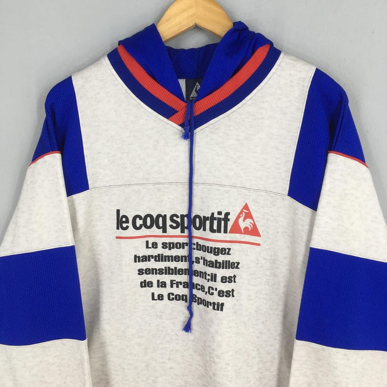 May include: A vintage Le Coq Sportif hoodie in white, blue, and red. The hoodie features the brand's logo and French text. The design includes blue panels on the sleeves and a blue and red striped neckline. The hoodie is hanging on a wooden hanger.