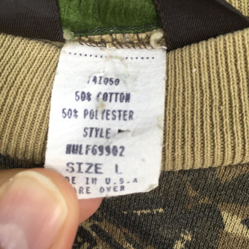 Op de afbeelding: Close-up van een kledinglabel met de tekst "50% COTTON, 50% POLYESTER, SIZE L, MADE IN U.S.A." Het label is wit met zwarte tekst en bevestigd aan een stof met camouflagepatroon.