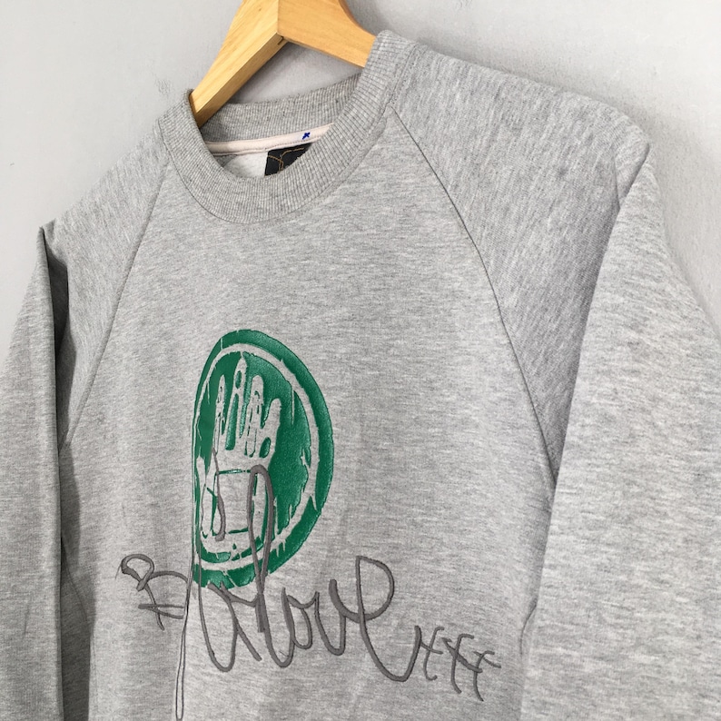 May include: A heather gray sweatshirt with a crew neck and long sleeves. The front features a green circular graphic with a handprint design and the word "Glove" in cursive script. The sweatshirt is hanging on a wooden hanger.