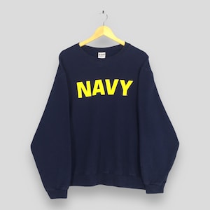 Könnte beinhalten: Marineblaues Sweatshirt mit dem Wort "NAVY" in großen gelben Blockbuchstaben auf der Brust. Das Sweatshirt hängt an einem Holzbügel vor neutralem Hintergrund. Das Kleidungsstück hat lange Ärmel und einen Rundhalsausschnitt.