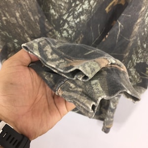 Puede incluir: Manga de sudadera con estampado de camuflaje, con una mezcla de tonos marrones, verdes y grises. La textura de la tela es visible, con el pu&ntilde;o doblado. El dise&ntilde;o sugiere un tema de ropa de caza o actividades al aire libre.