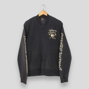 May include: Black zip-up jacket with the text "Vanson Leathers" and a skull and crossbones graphic. The sleeves feature the text "Vanson Leathers" in a gold color. The jacket has two front pockets and a striped lining.