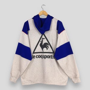 May include: A gray sweatshirt with blue and white accents, featuring a large black triangle with a rooster logo and the text "le coq sportif." The long-sleeved top has a blue hood and is hanging on a wooden hanger.