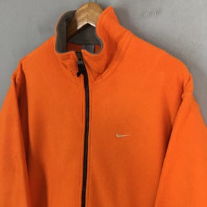 May include: Vibrant orange Nike fleece jacket with a high collar and a full-length black zip. The jacket has a small white Nike swoosh logo on the chest. The fleece material looks soft and warm, suitable for cooler weather.