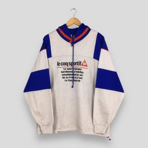 May include: A gray Le Coq Sportif hoodie with blue and red accents. The hoodie features a drawstring hood, long sleeves, and the brand's logo and text in black and red. The design includes blue panels on the sleeves.
