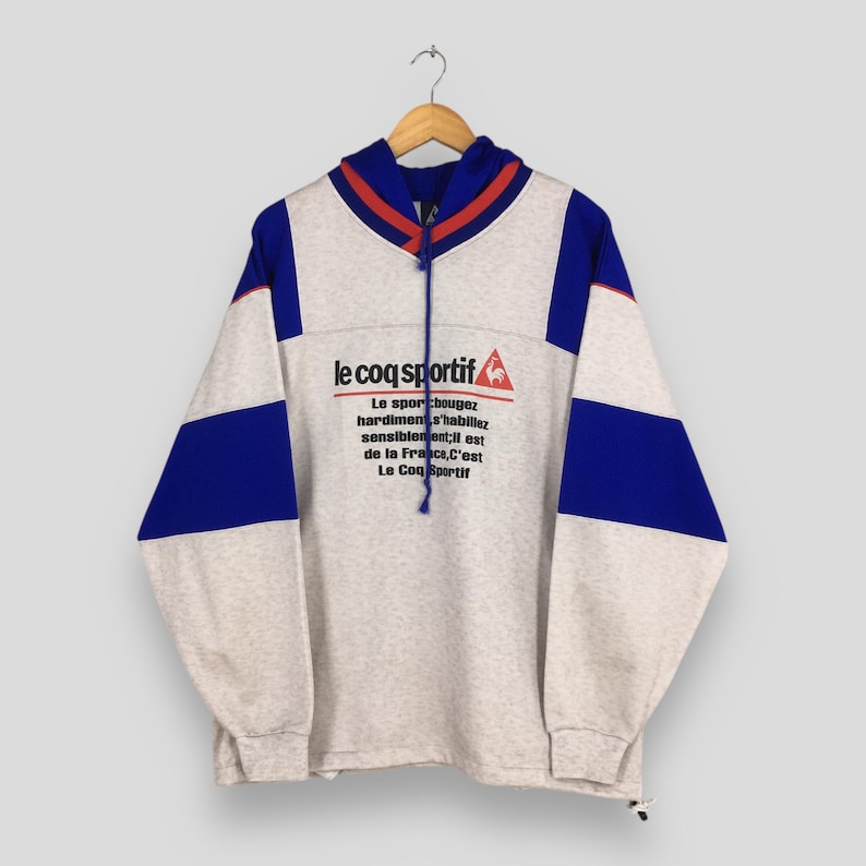 May include: A gray Le Coq Sportif hoodie with blue and red accents. The hoodie features a drawstring hood, long sleeves, and the brand's logo and text in black and red. The design includes blue panels on the sleeves.