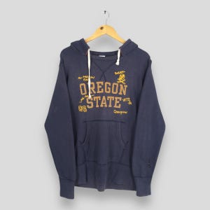Puede incluir: Sudadera con capucha azul marino con la inscripción "Oregon State" en letras doradas. La sudadera tiene un bolsillo de canguro y cordones blancos. El diseño incluye el año "1972" y un gráfico de calavera y huesos cruzados.